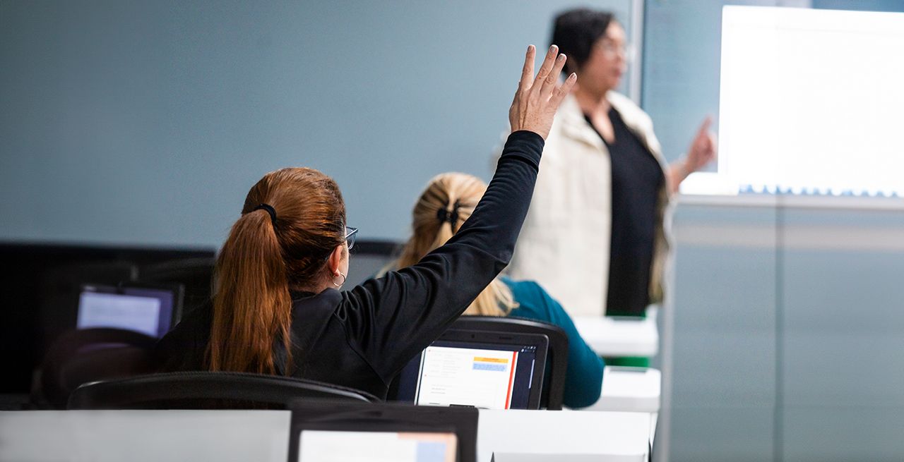 Student raising her hand in a computer lab, teacher at whiteboard in the background