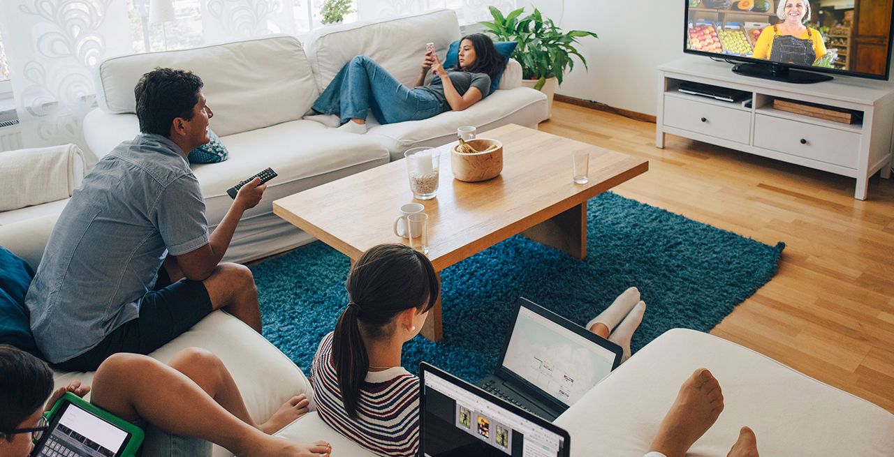 Family using various devices - laptops, phones, tablets - in their living room with the TV on